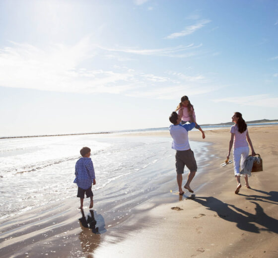 Rear View Of Family Walking Along Beach With Picnic Basket