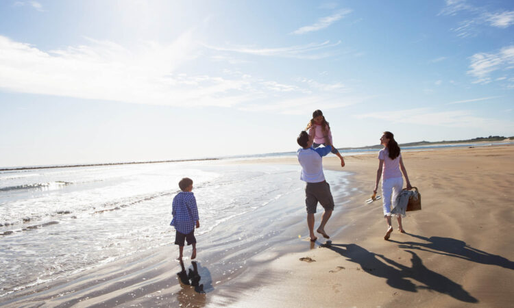 Rear View Of Family Walking Along Beach With Picnic Basket
